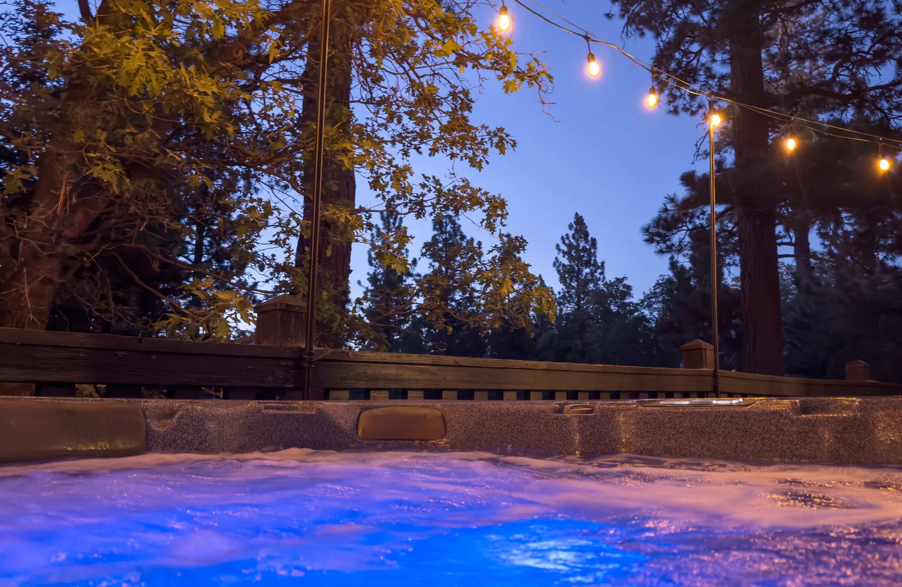 Private hot tub at Pine Grove Cabin beneath glowing string lights and tall pine trees at dusk